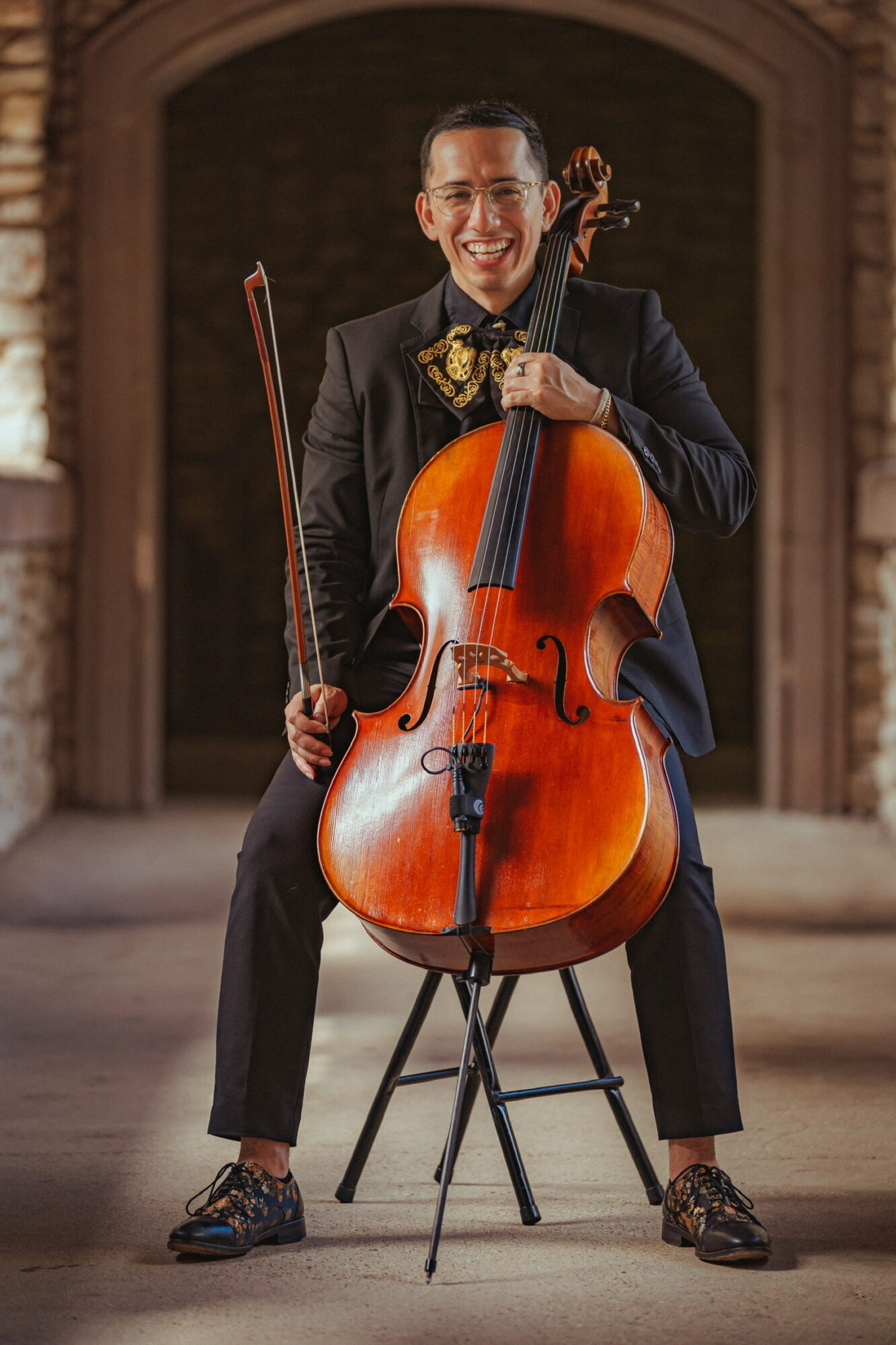 Man in formal attire smiling while holding a cello, sitting on a chair in front of an arched background.