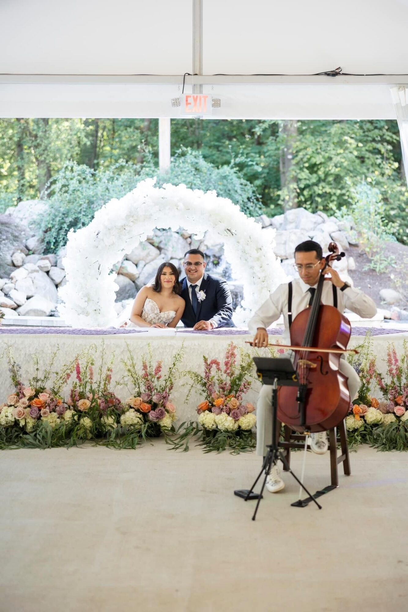 Wedding couple and musician playing cello indoors with a fountain and greenery outside window, decorated with flowers.