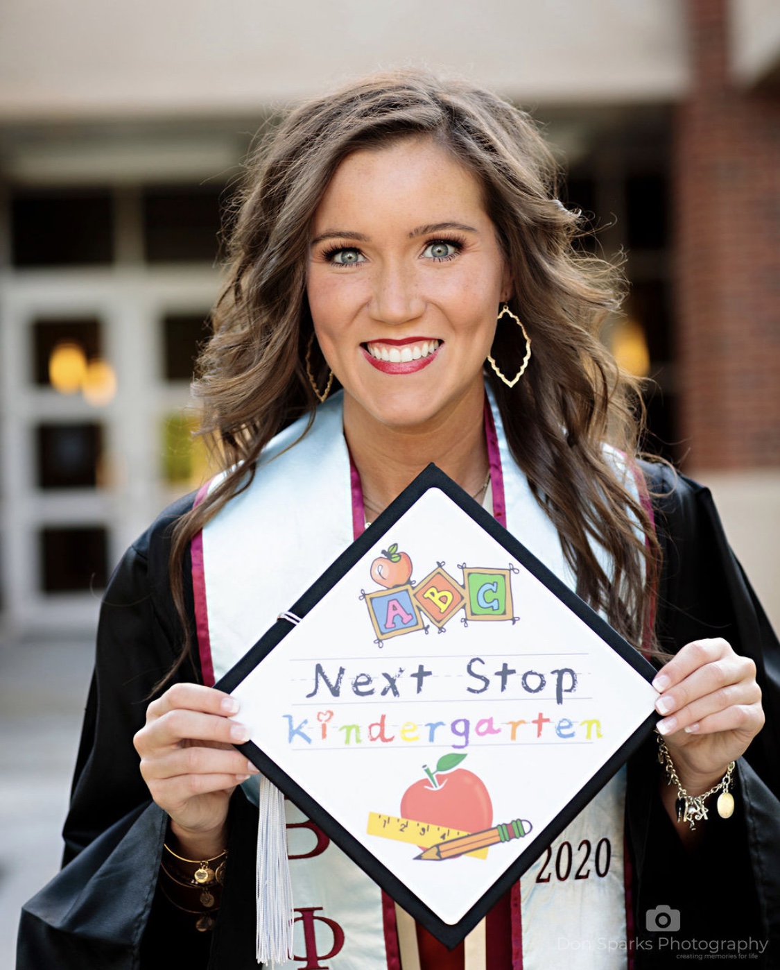 Young woman smiling holding a kindergarten graduation cap with colorful letters and drawings, outdoors background.