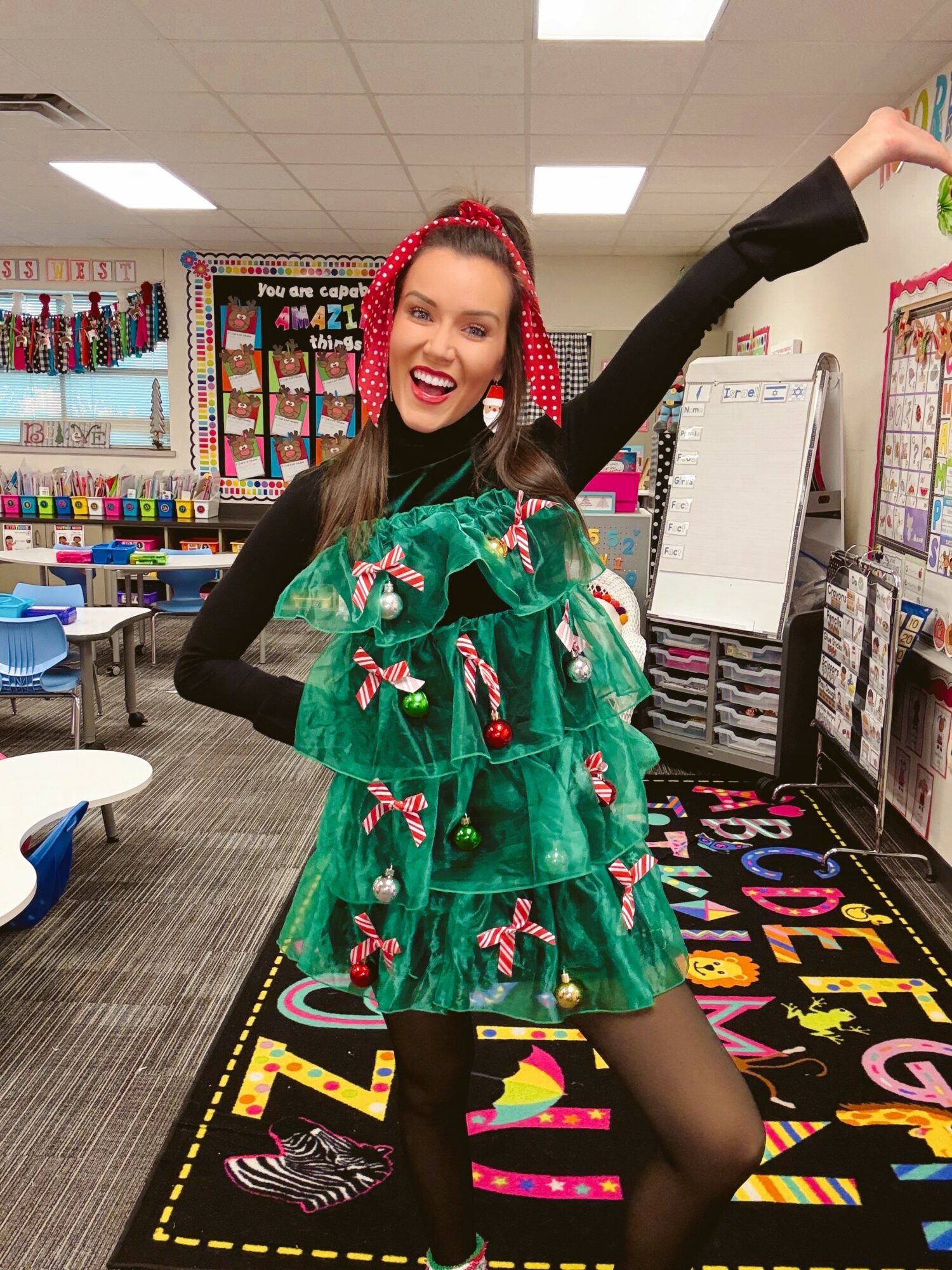 Woman in festive costume with green ruffles, red headband, smiling, in classroom with colorful decorations and alphabet rug.