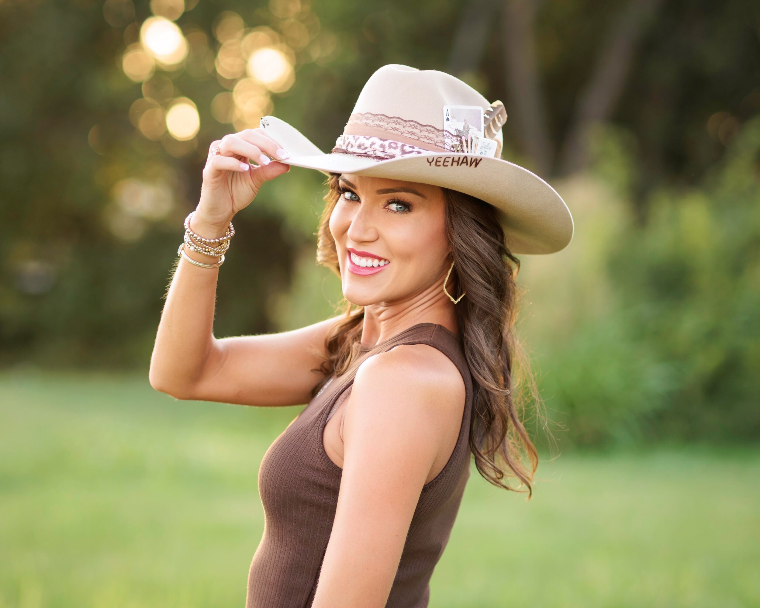 Woman smiling outdoors, wearing a wide-brimmed hat and sleeveless top, holding the hat's brim with one hand.