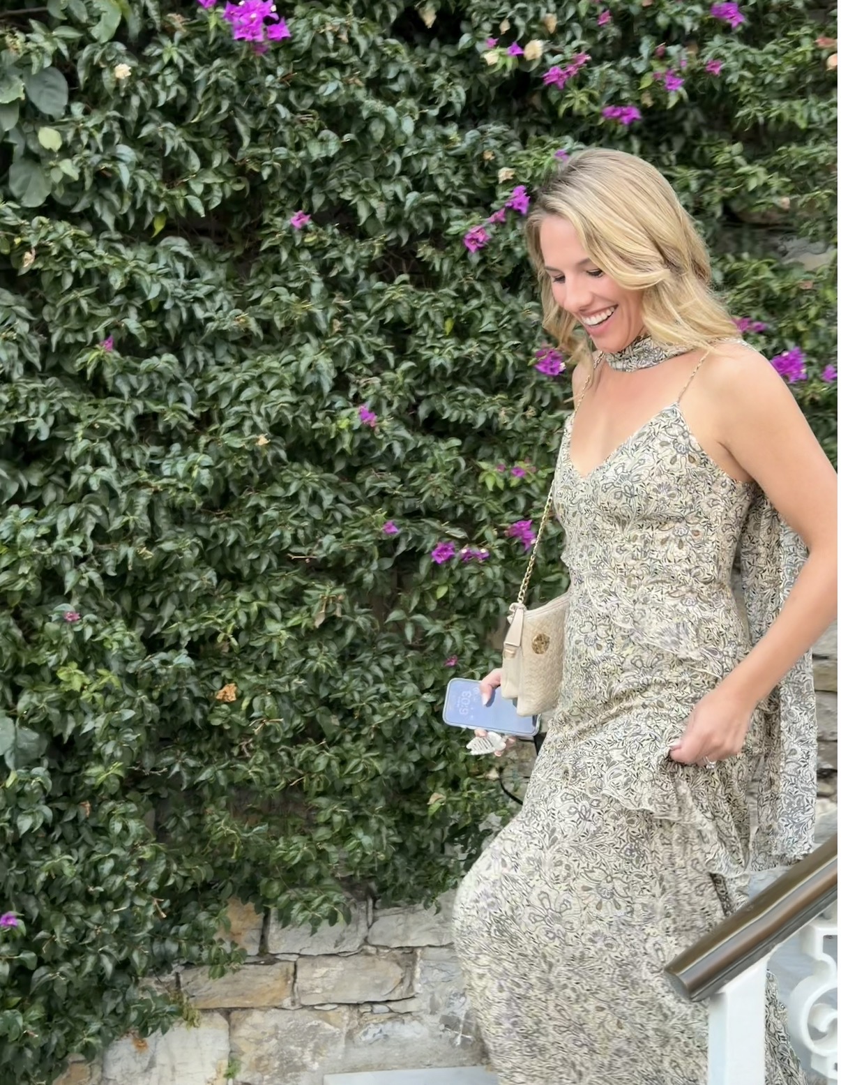 Woman in floral dress smiling outdoors with green bushes and purple flowers behind her.