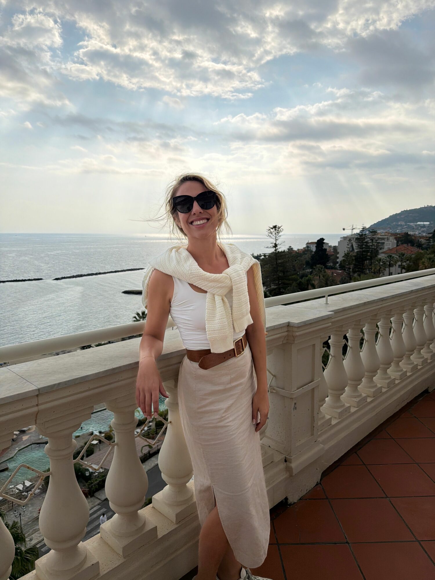Woman in white outfit and sunglasses standing on balcony with water and sky in background.