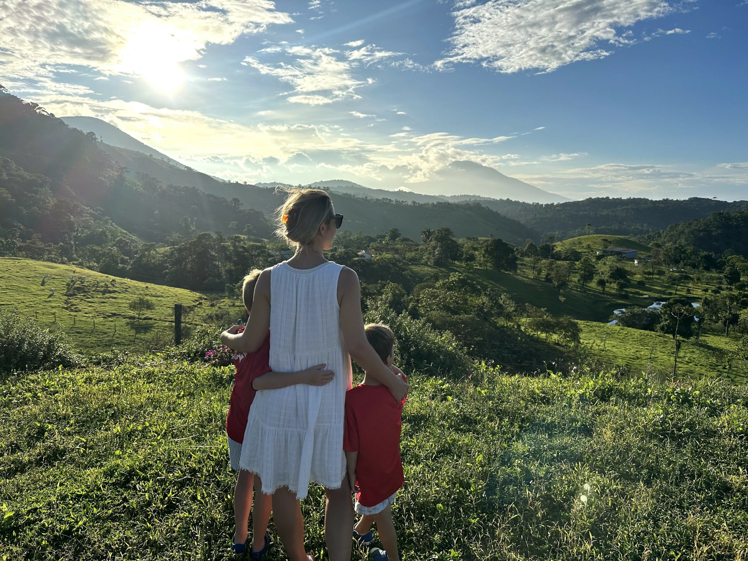 Woman and child standing on a grassy hill overlooking a landscape with mountains and a partly cloudy sky.