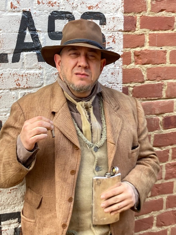 Man in vintage clothing holding a drink, standing against a brick wall with partially visible text.
