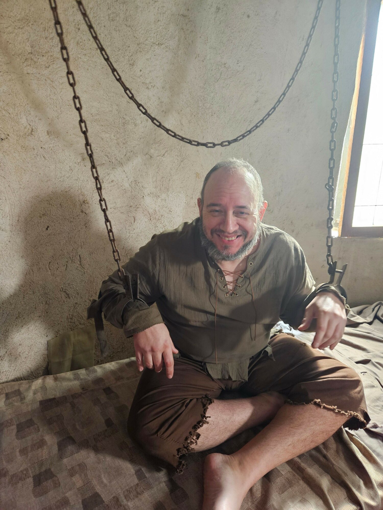An elderly man with gray hair and a beard sitting on a bed, smiling, with a chain hanging on the wall behind him.