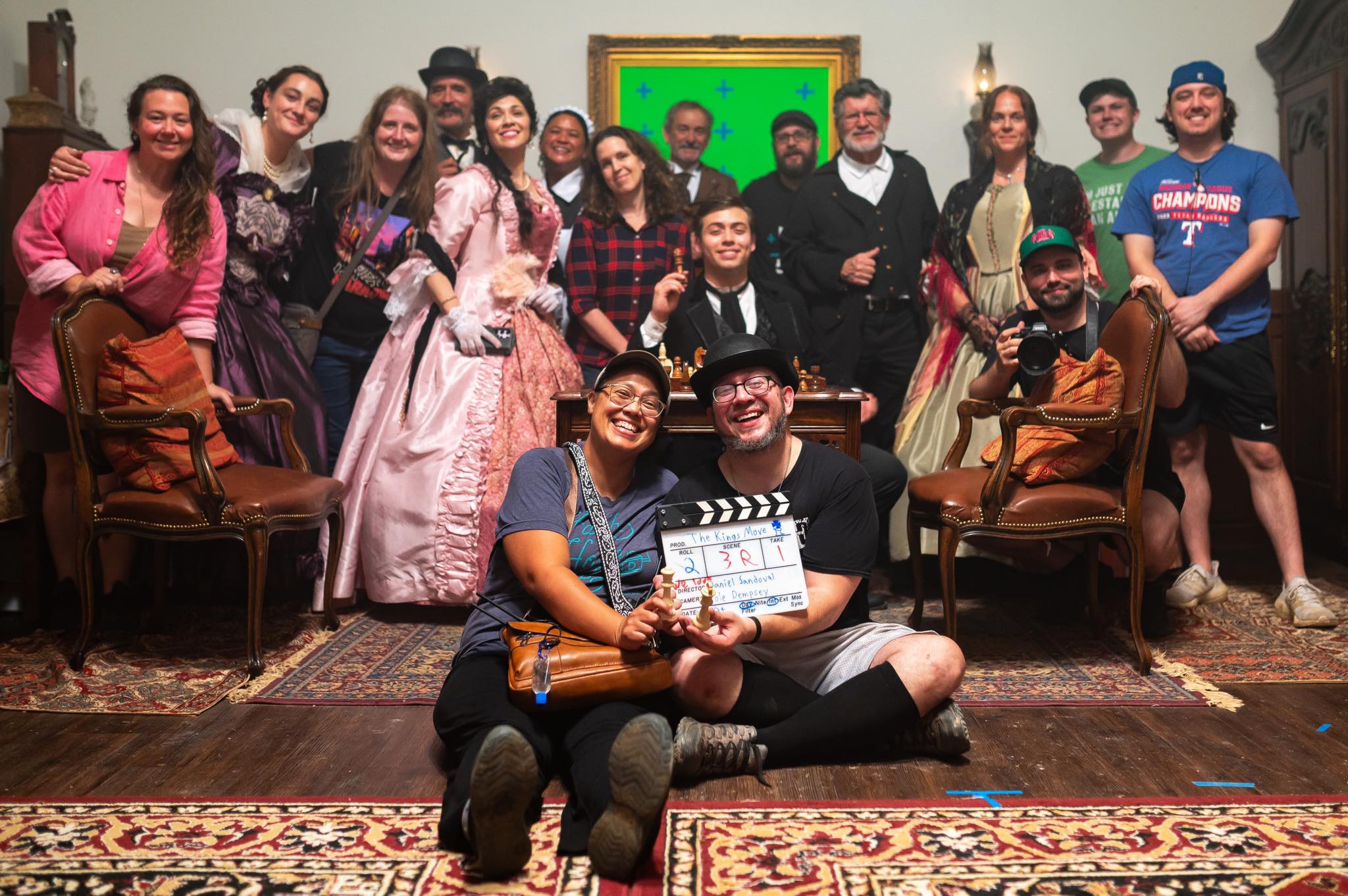 Group of people posing indoors, some in costumes, with chairs and a framed picture on the wall behind them.