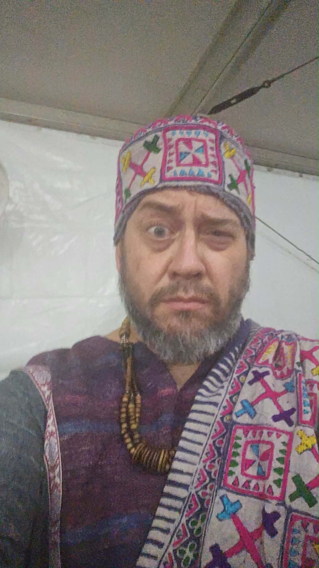 Man wearing a colorful embroidered hat and a patterned garment, looking at the camera with a neutral expression.