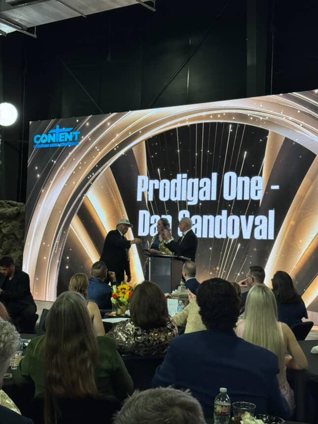 People seated at tables watching a presentation on a large screen with the text 'Prodigal One - Dan Candoval'.