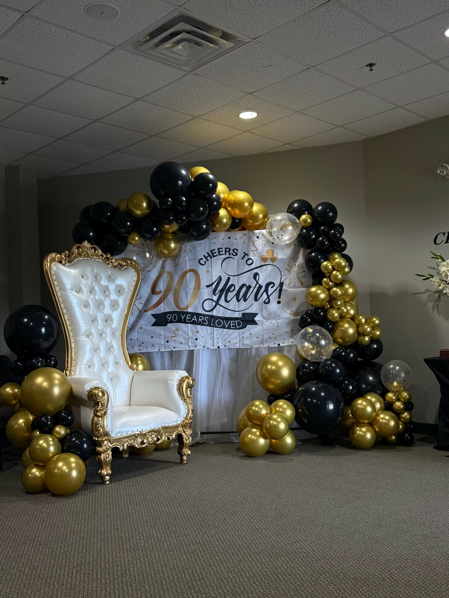 Decorative backdrop with black and gold balloons, a white ornate chair, and a banner reading 'Welcome' in a room.