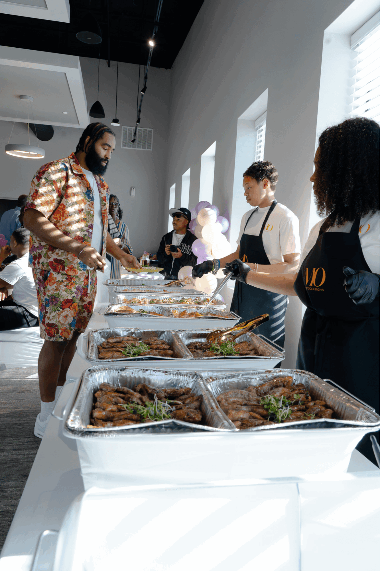 Person in colorful clothing serving food at a buffet table with others in aprons, in a bright room with large windows.