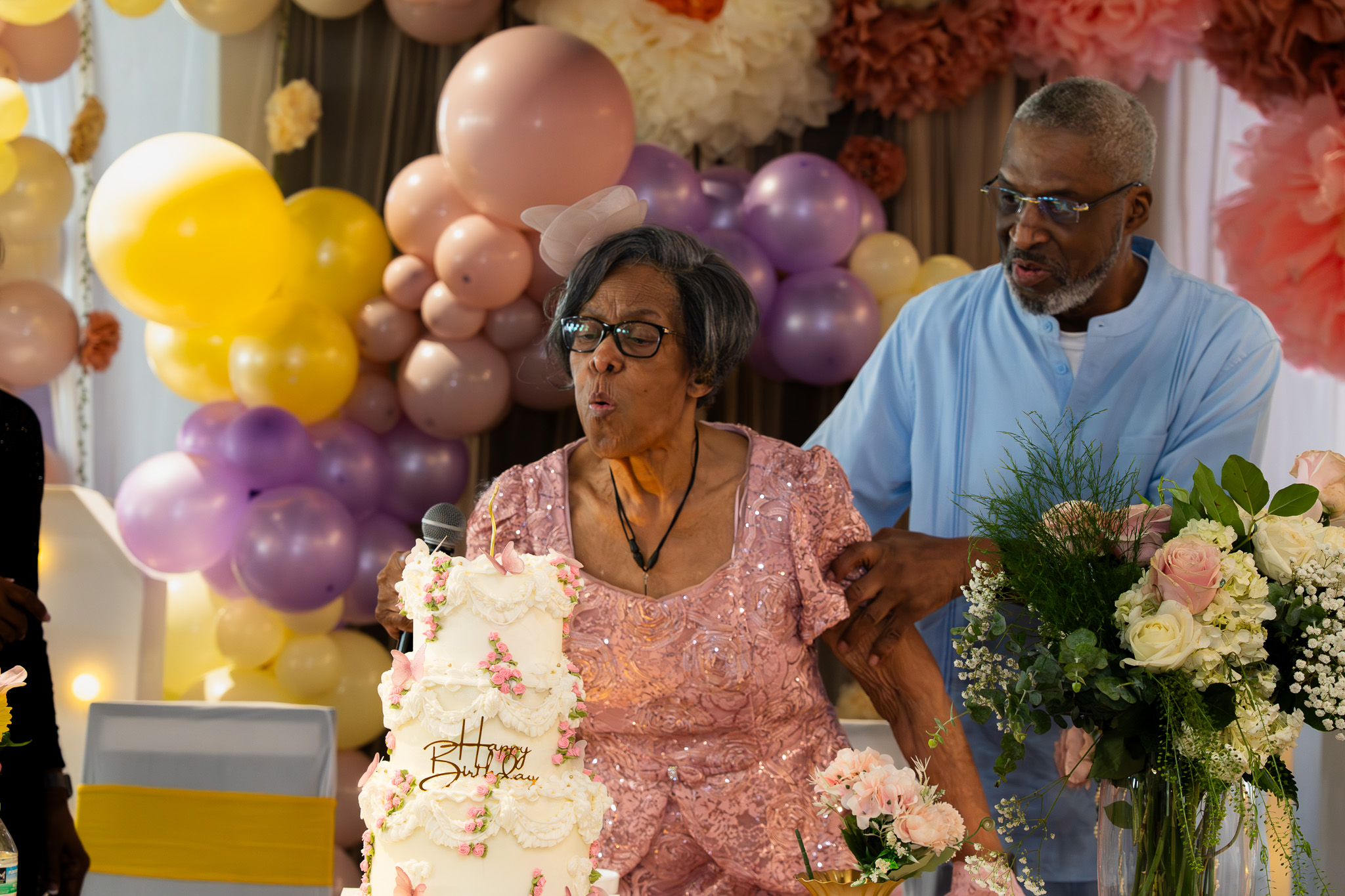 Woman and man at a celebration with balloons and flowers, woman in pink dress, man in blue shirt, woman holding a cake