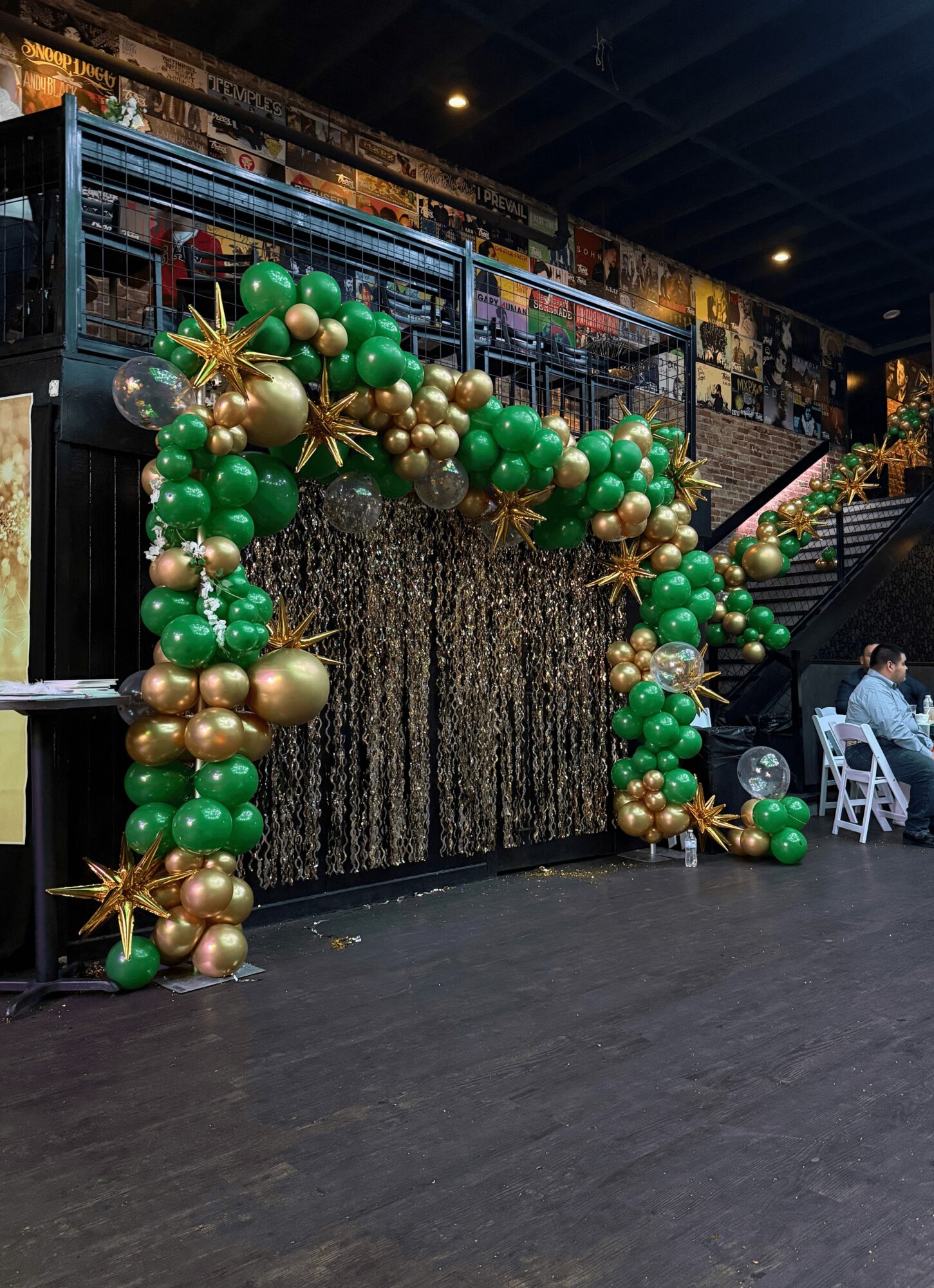 Decorative arch made of green, gold, and silver balloons with star-shaped accents, set against a textured backdrop in an indoor space.