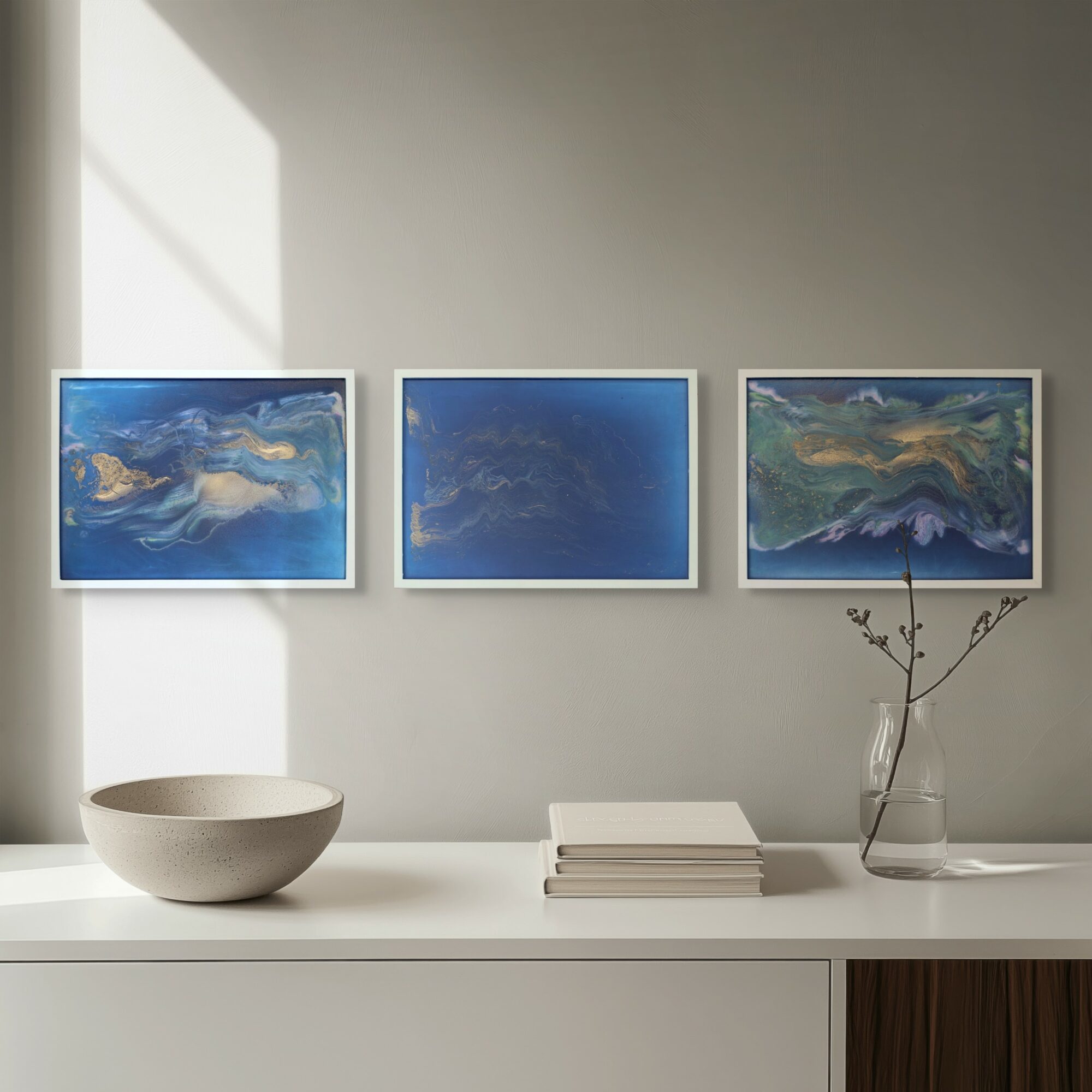 Three framed abstract paintings hang on a light gray wall above a white sideboard, with a bowl, books, and a vase with branches.