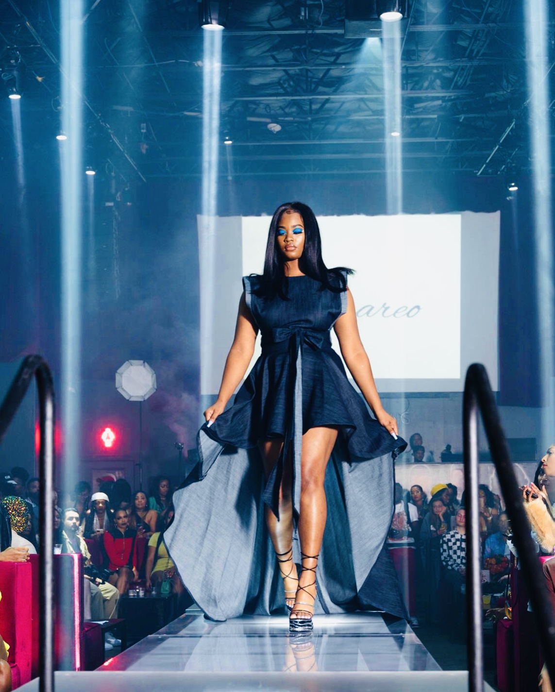 Woman walking on runway in a dark blue dress with high heels, audience seated on both sides, stage lights overhead.