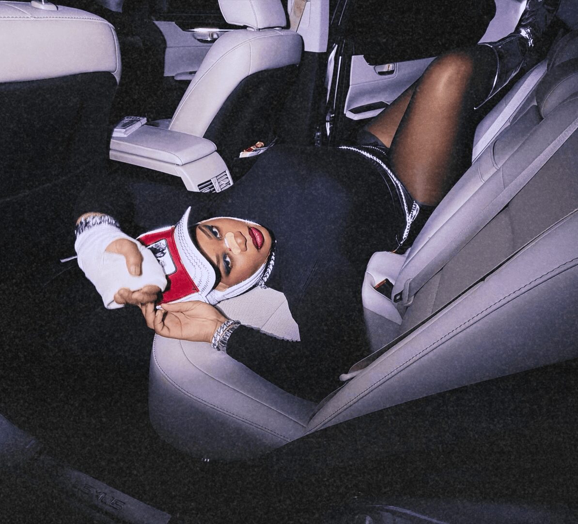 Person lying on car seat with legs up, wearing black outfit and a headscarf, holding a phone, inside a vehicle.