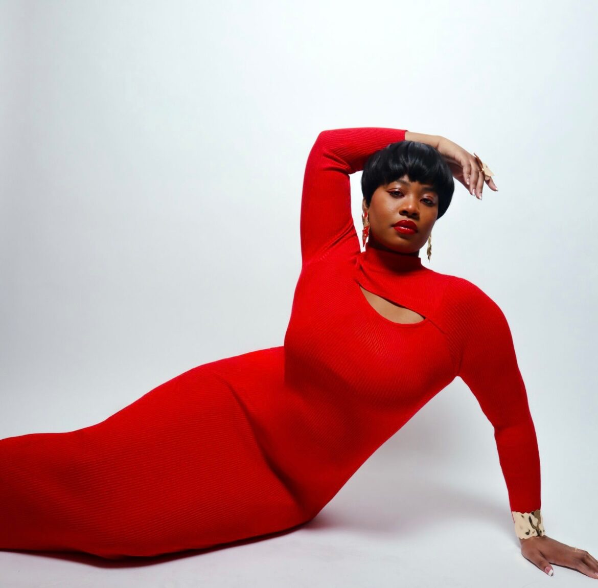 Woman in red dress lying on the floor with her arm raised above her head, against a plain white background.
