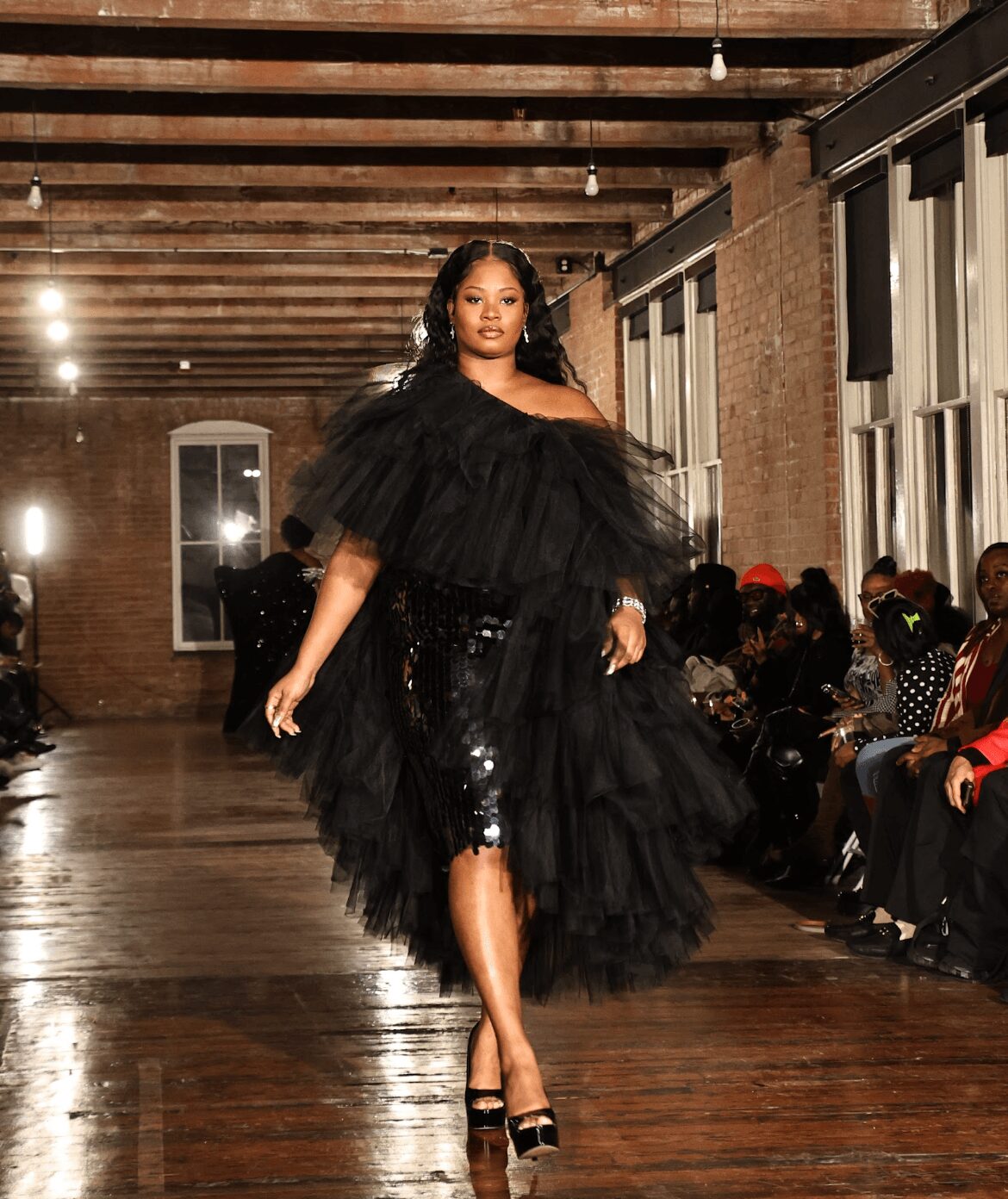 Model walking down runway in black ruffled dress with sequined skirt, audience seated on sides, industrial-style venue.