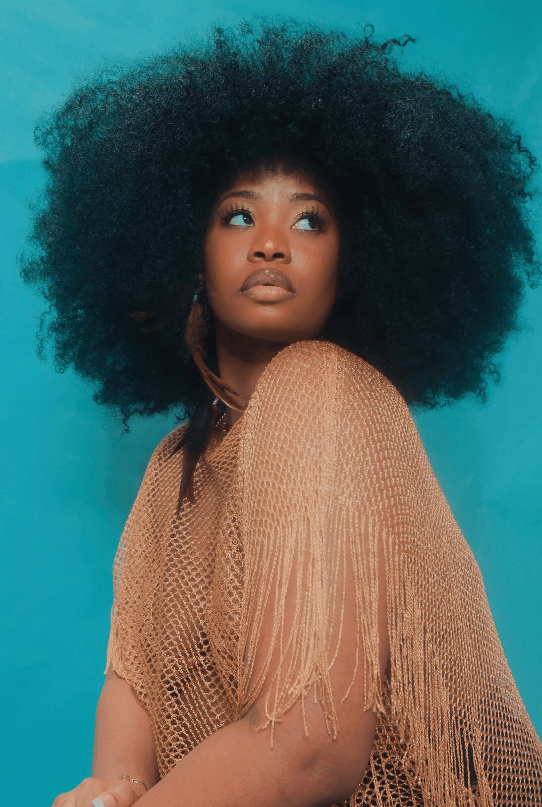 Portrait of a woman with large, curly hair wearing a tan fringed top against a blue background.