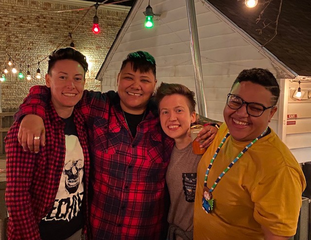 Four people smiling and posing together outdoors at night, with string lights overhead and a white structure behind them.