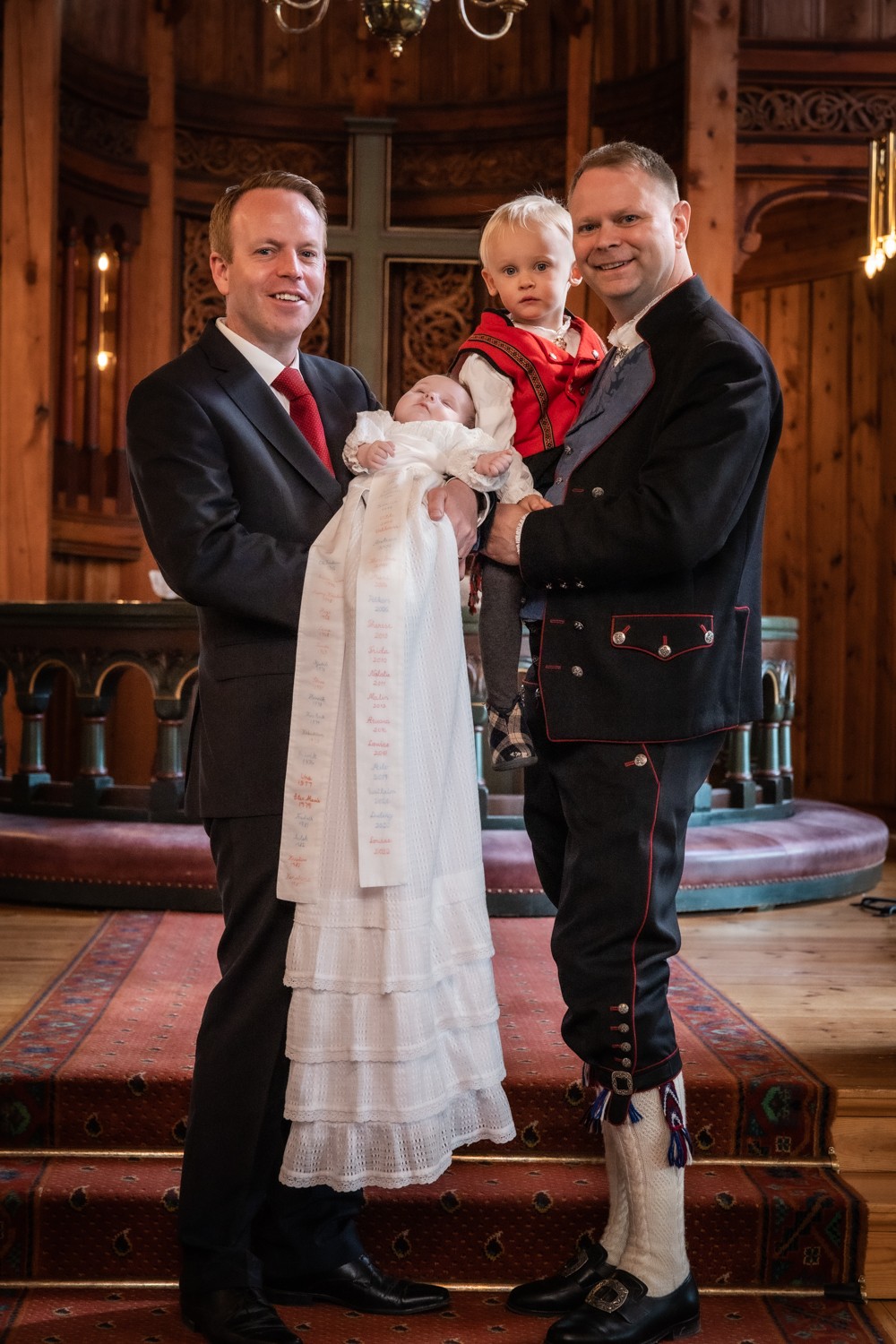 Two men in formal attire holding a child and a white dress, standing on a wooden staircase inside a decorated room.