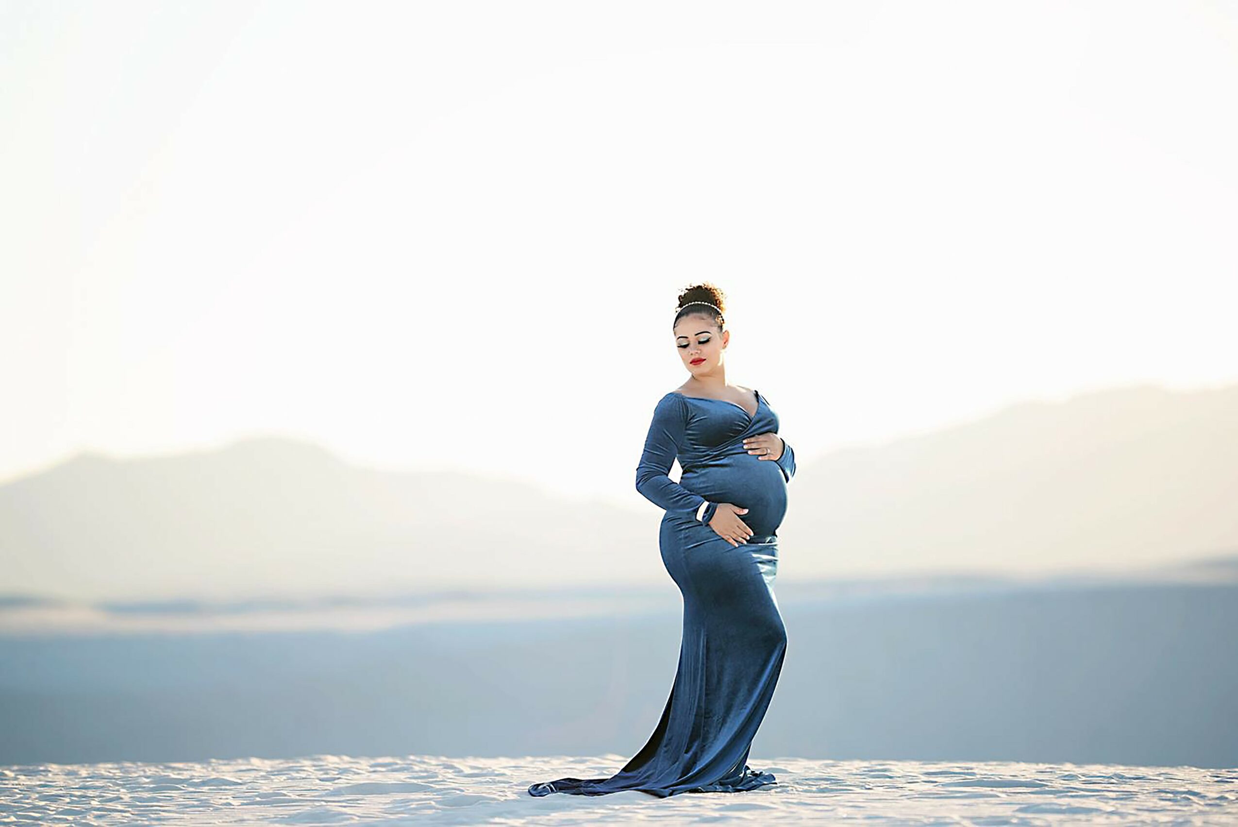 Pregnant woman in a long blue dress standing outdoors with a blurred landscape background.