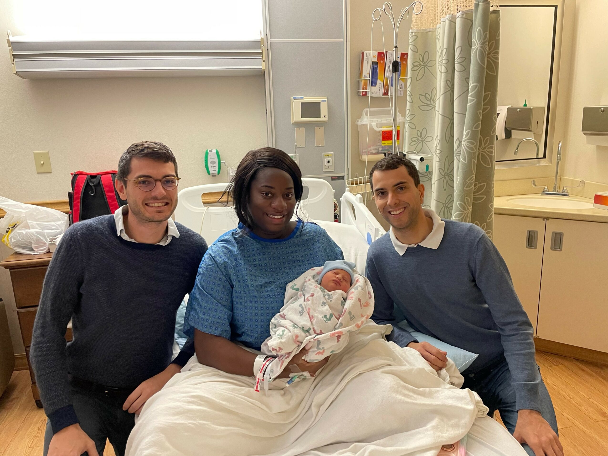 Three people, including a woman holding a newborn, sit on a hospital bed in a medical room.