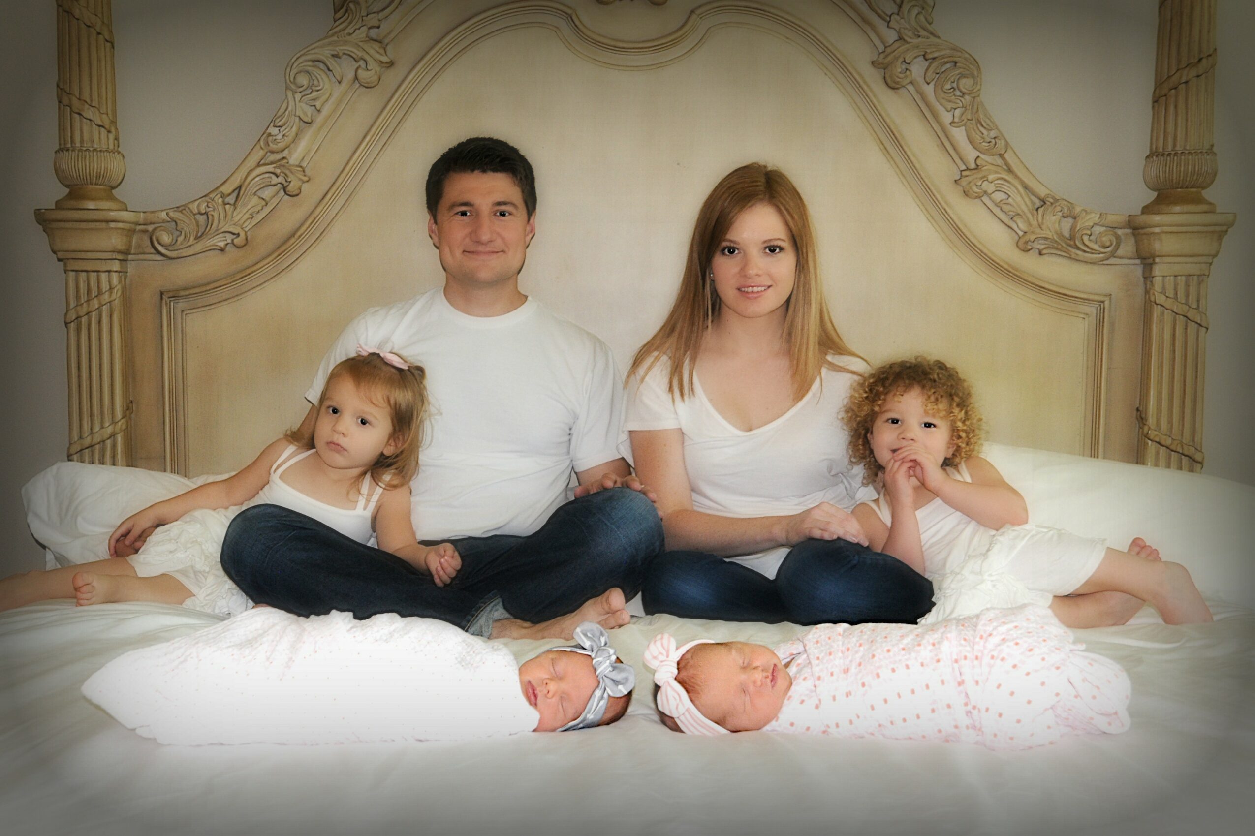 Family of four sitting on a bed with a decorative headboard, two children lying in front, parents behind, all smiling.