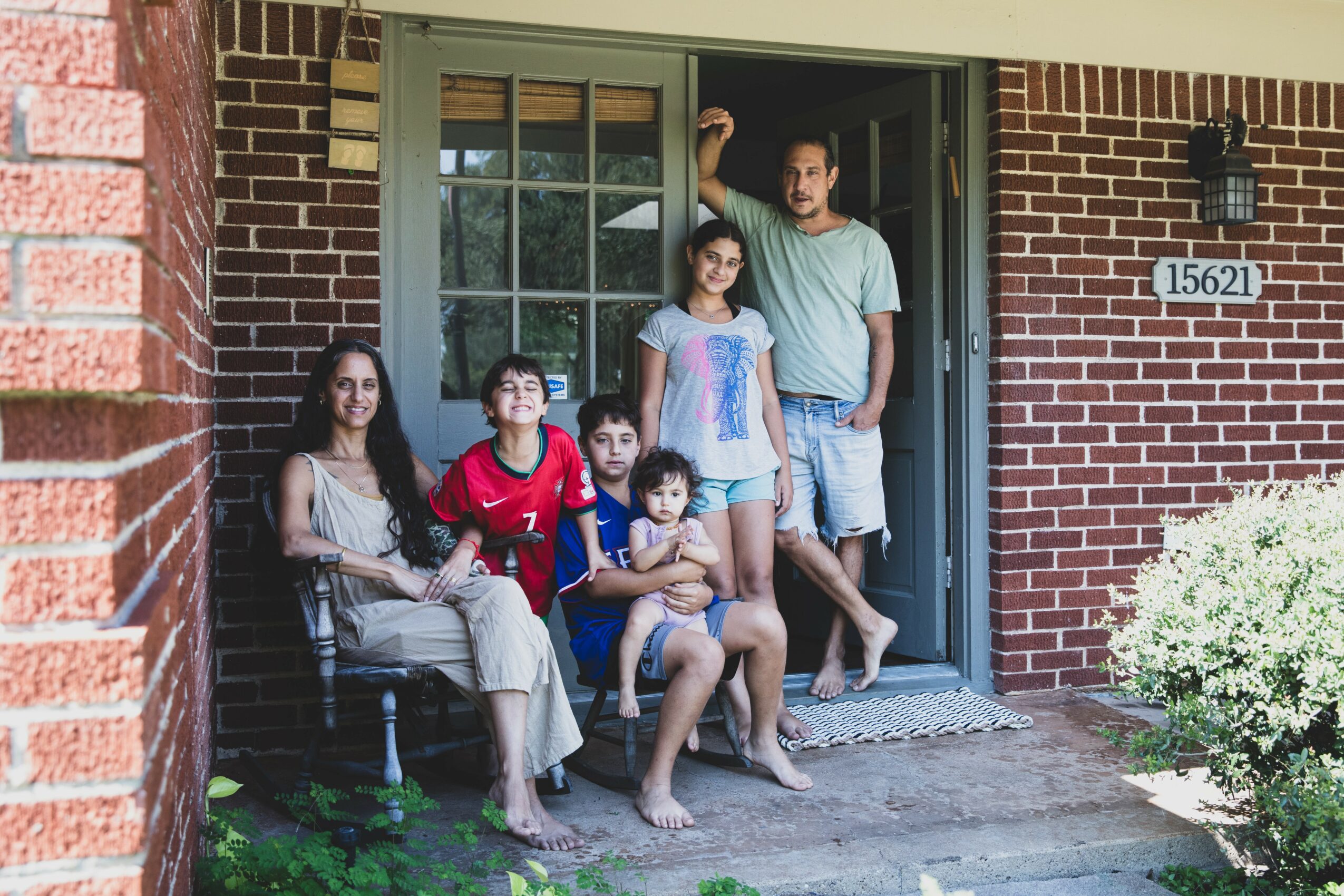 Family of six sitting and standing on porch in front of brick house, with a man standing at the door, children sitting on steps.
