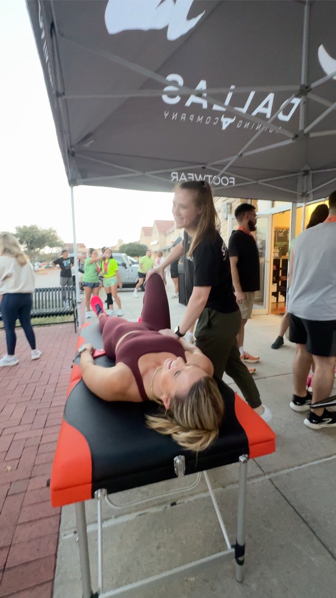 Person lying on a massage table outdoors, receiving a massage from a therapist, with people and a tent in the background.