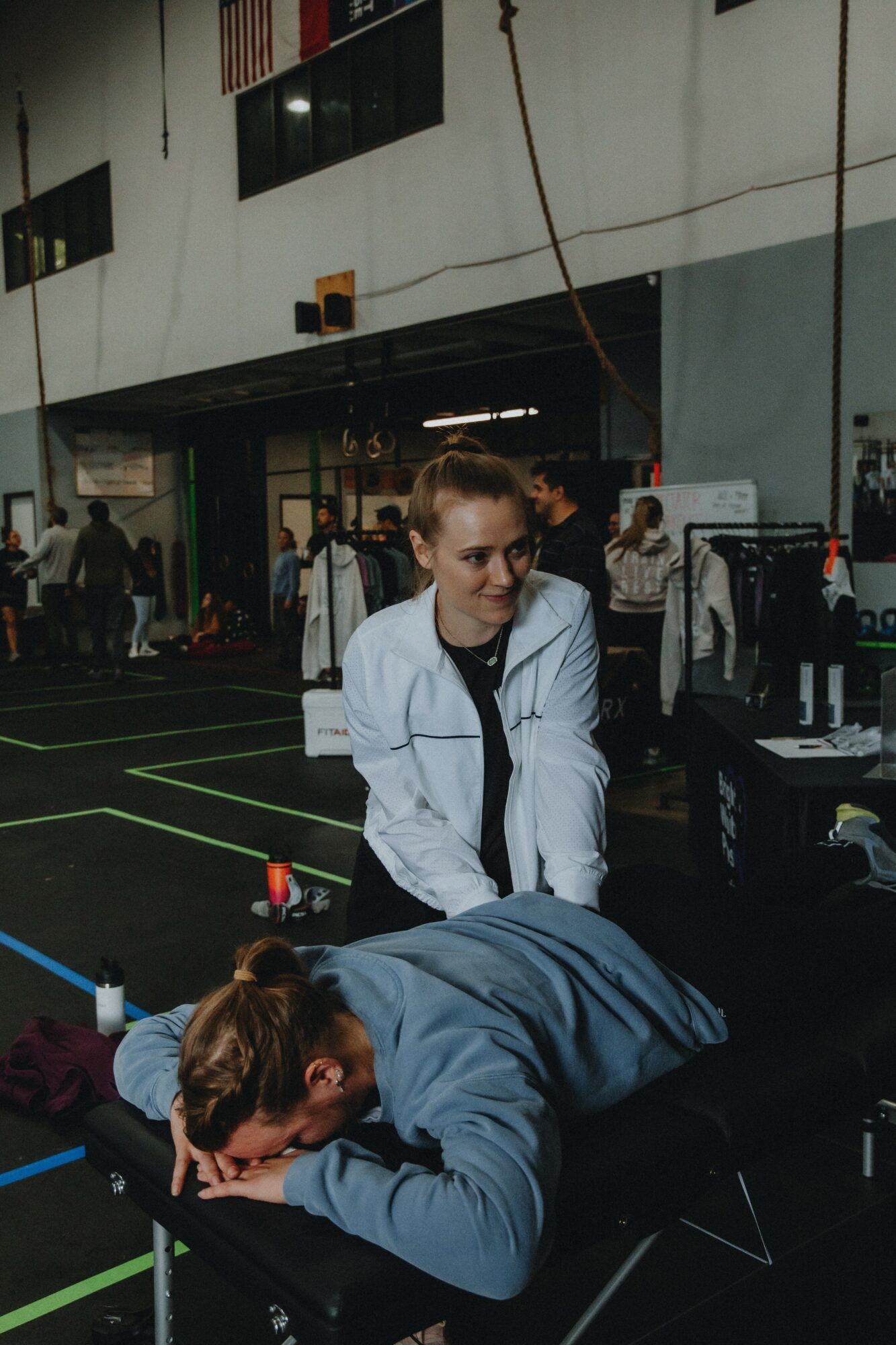 Two women in a gym or training facility, one standing and the other lying on a table, with equipment and people in the background.