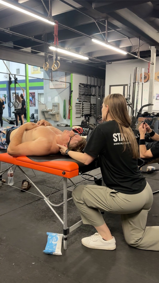 Person receiving a massage on an orange table in a gym, with a therapist kneeling beside them, gym equipment visible in background.