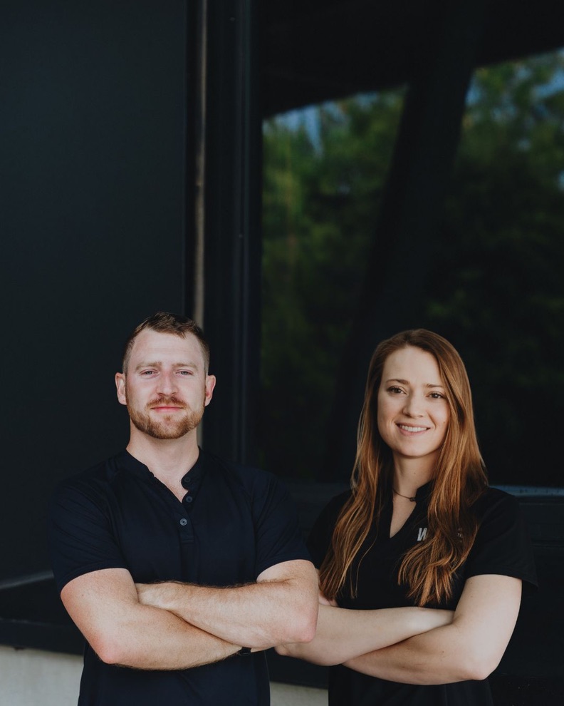 A man and woman stand side by side outdoors, smiling, with trees and a building in the background.