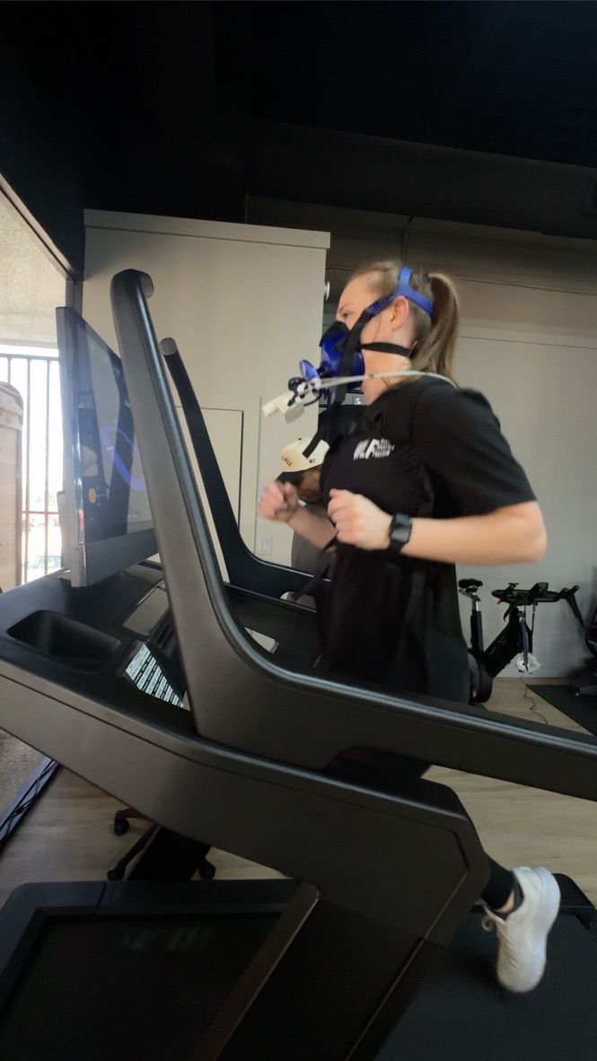 Person wearing a mask and glasses using a treadmill in a gym, with a bicycle in the background.
