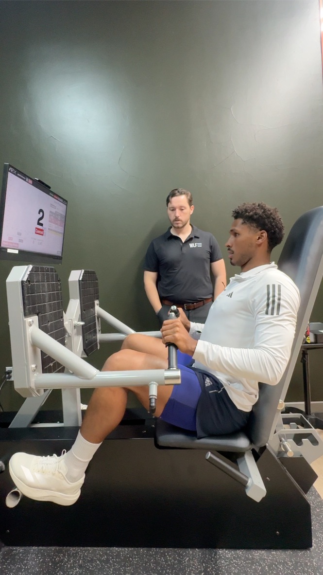 Young man seated on exercise machine with a trainer standing nearby, in a gym setting.