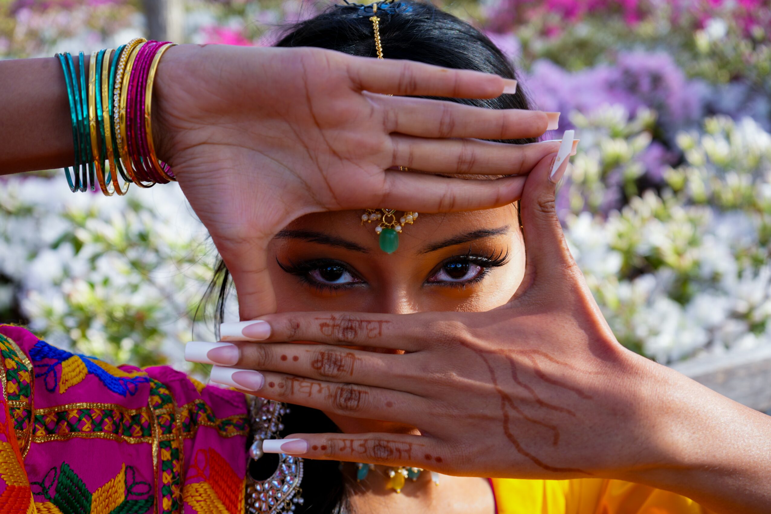 Person with hands framing face, wearing colorful jewelry and traditional attire, with a floral background.