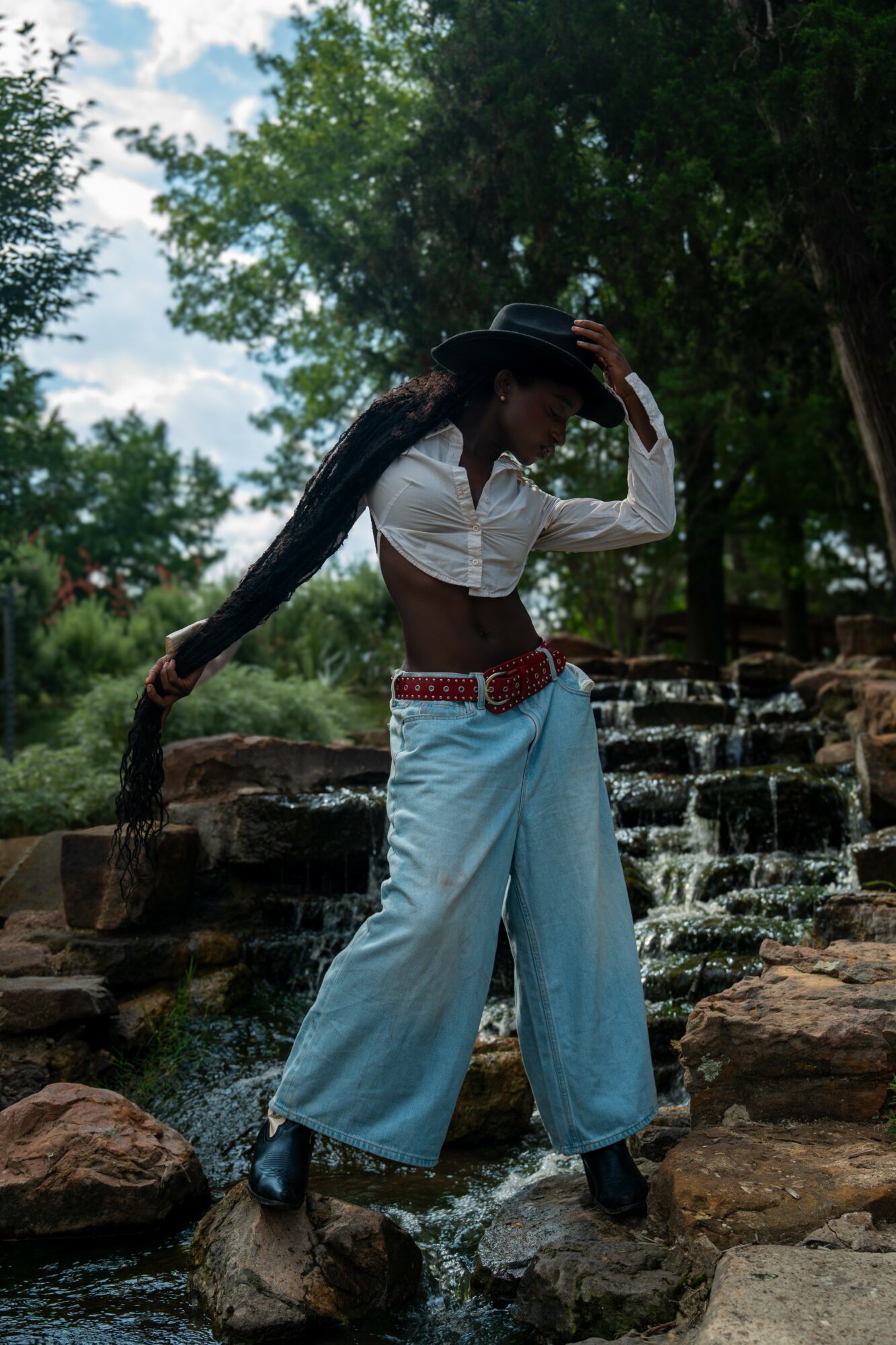 Person standing on rocks in a stream, holding long hair, surrounded by trees and water, outdoors.