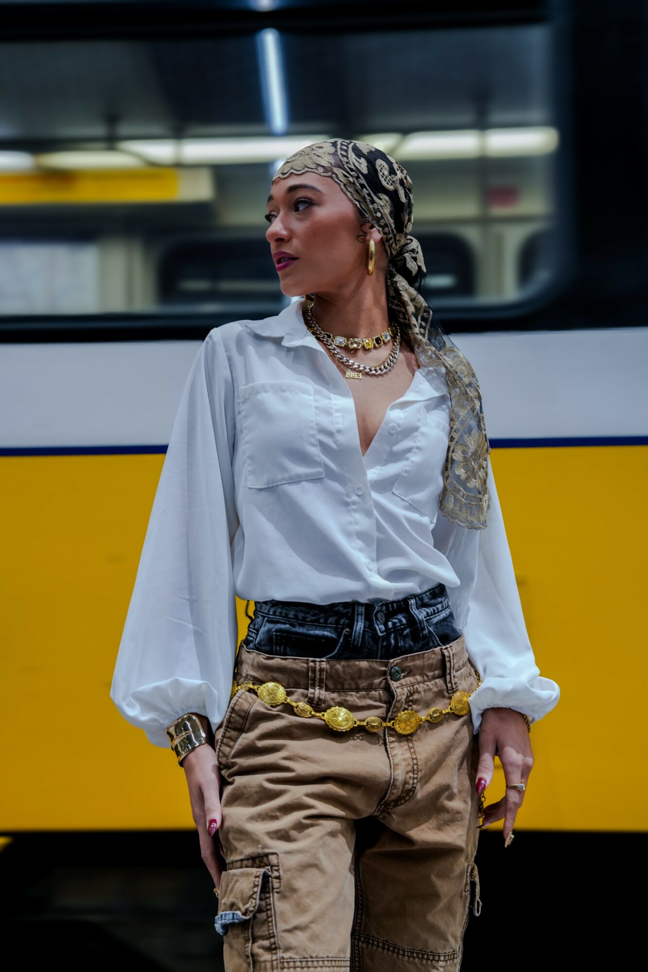 Woman standing in front of a yellow and black vehicle, wearing a white blouse, beige cargo pants, and jewelry.