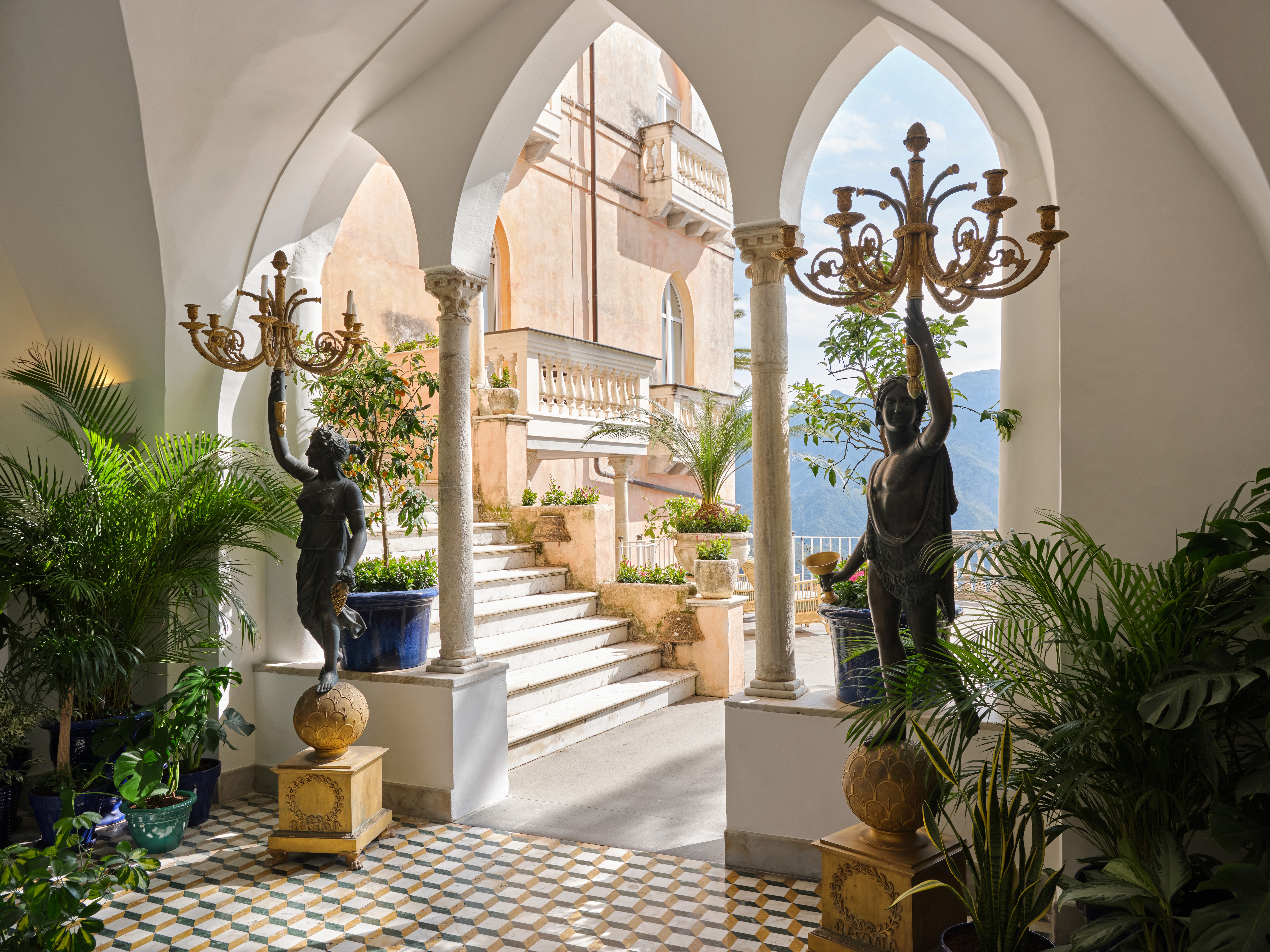 Elegant balcony with statues, potted plants, and a staircase leading to an outdoor view, under arched ceiling with chandeliers.