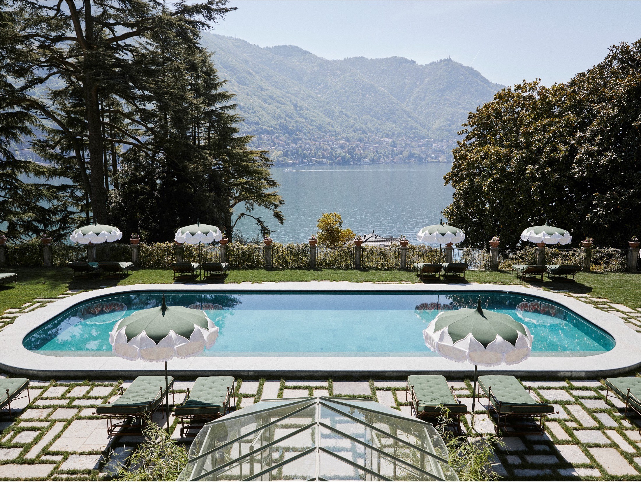 Swimming pool with umbrellas, surrounded by lounge chairs, overlooking a lake and mountains, trees on both sides, sunny day.