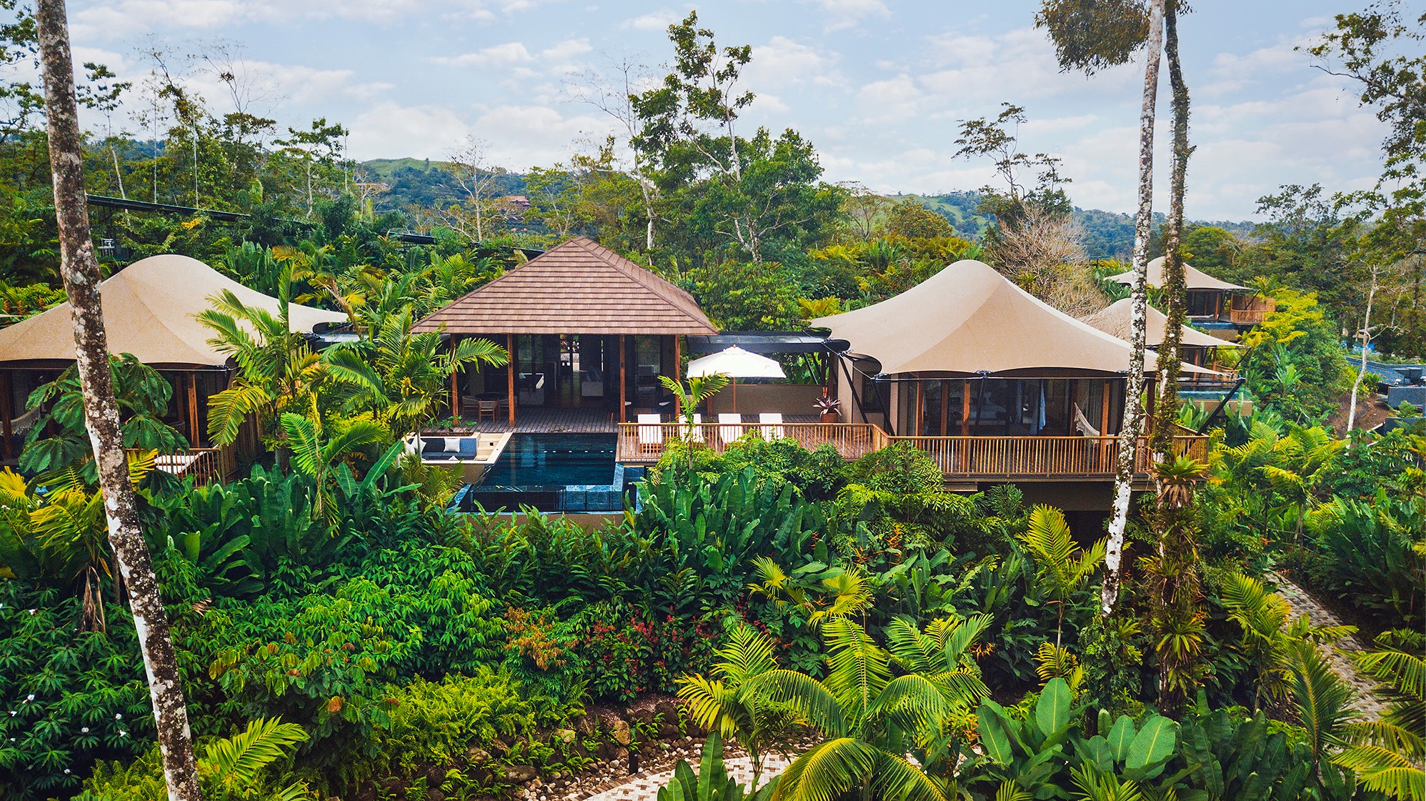 A tropical resort with multiple buildings and lush greenery, set against a backdrop of trees and a cloudy sky.