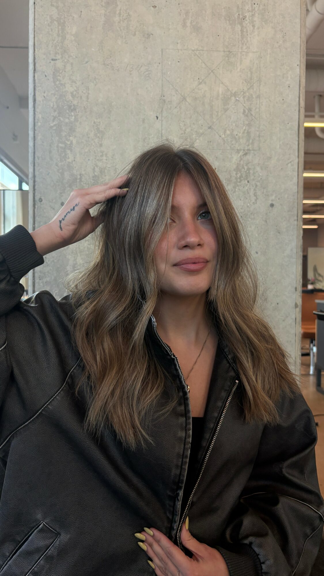 Woman with wavy hair wearing a black jacket, standing indoors near a concrete wall.