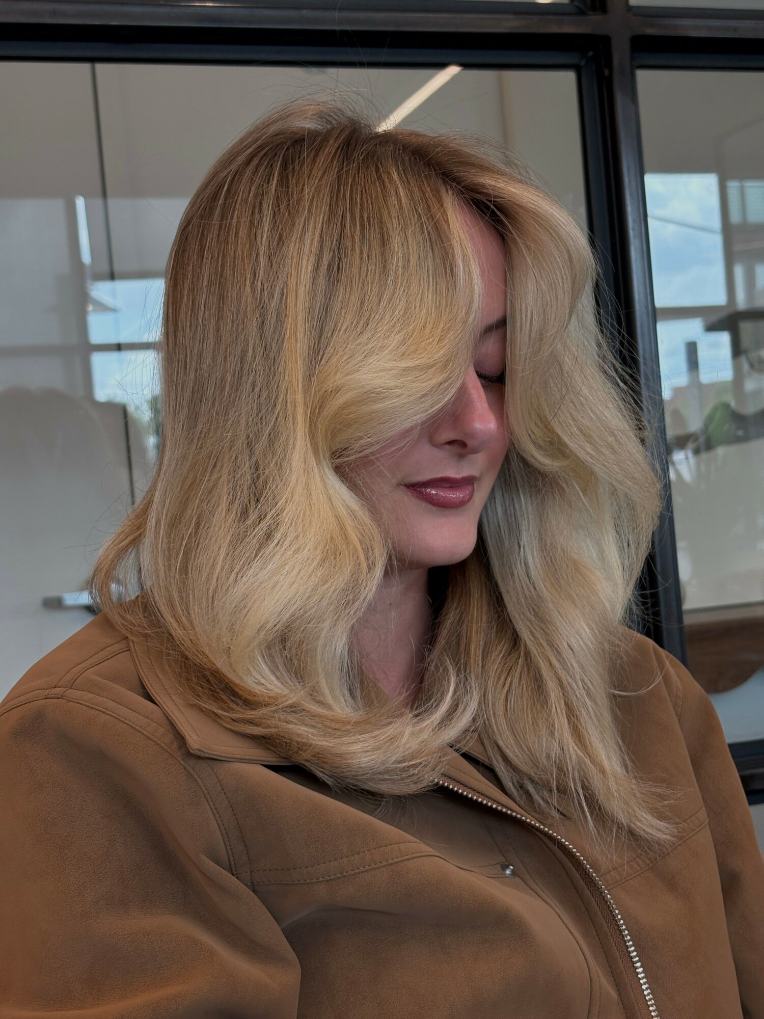 Woman with long, wavy blonde hair looking downward, wearing a brown jacket, indoors with large windows behind her.