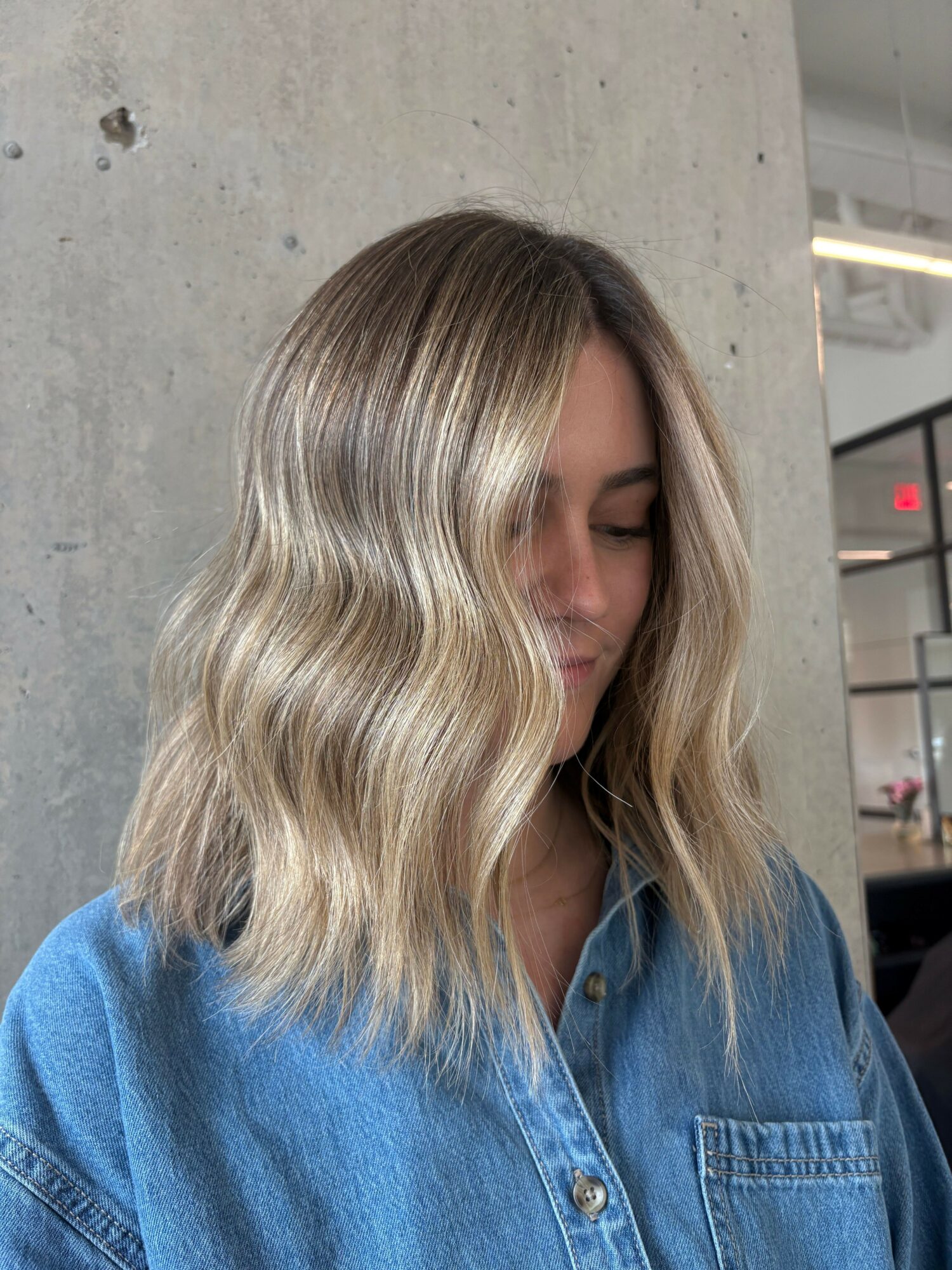 Side view of a woman with long, wavy blonde hair wearing a denim shirt, standing against a concrete wall.