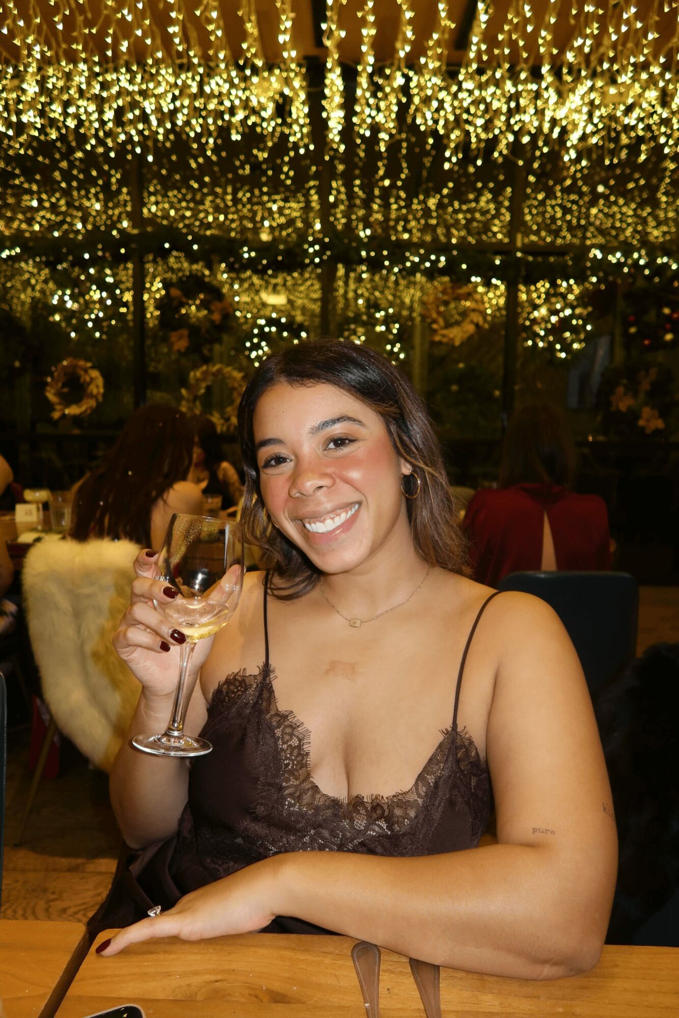Young woman smiling, holding a glass, sitting at a table in a decorated indoor setting with golden lights above.
