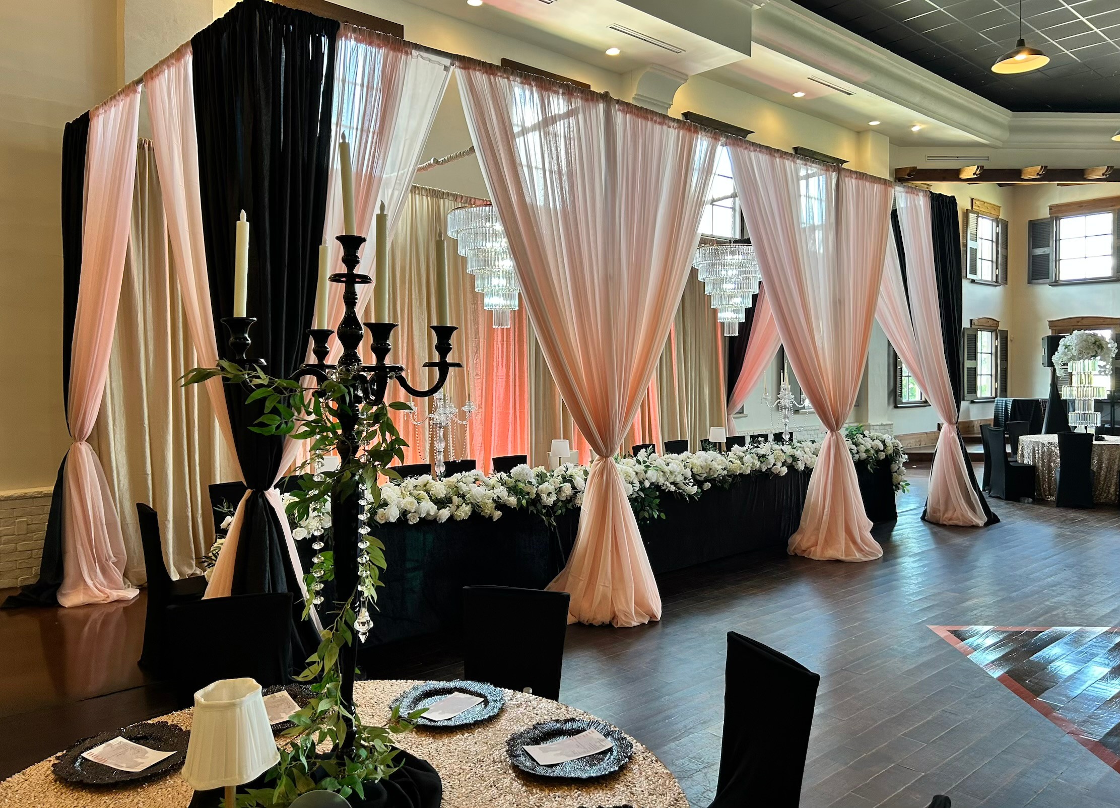Elegant event space with pink draped curtains, floral arrangements, and a candelabrum on a table, with a dark floor and high ceiling.