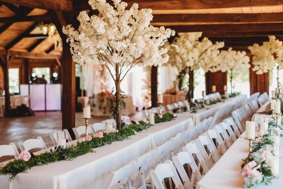 Long banquet table decorated with white flowers, candles, and greenery in a wooden venue with large floral arrangements overhead.