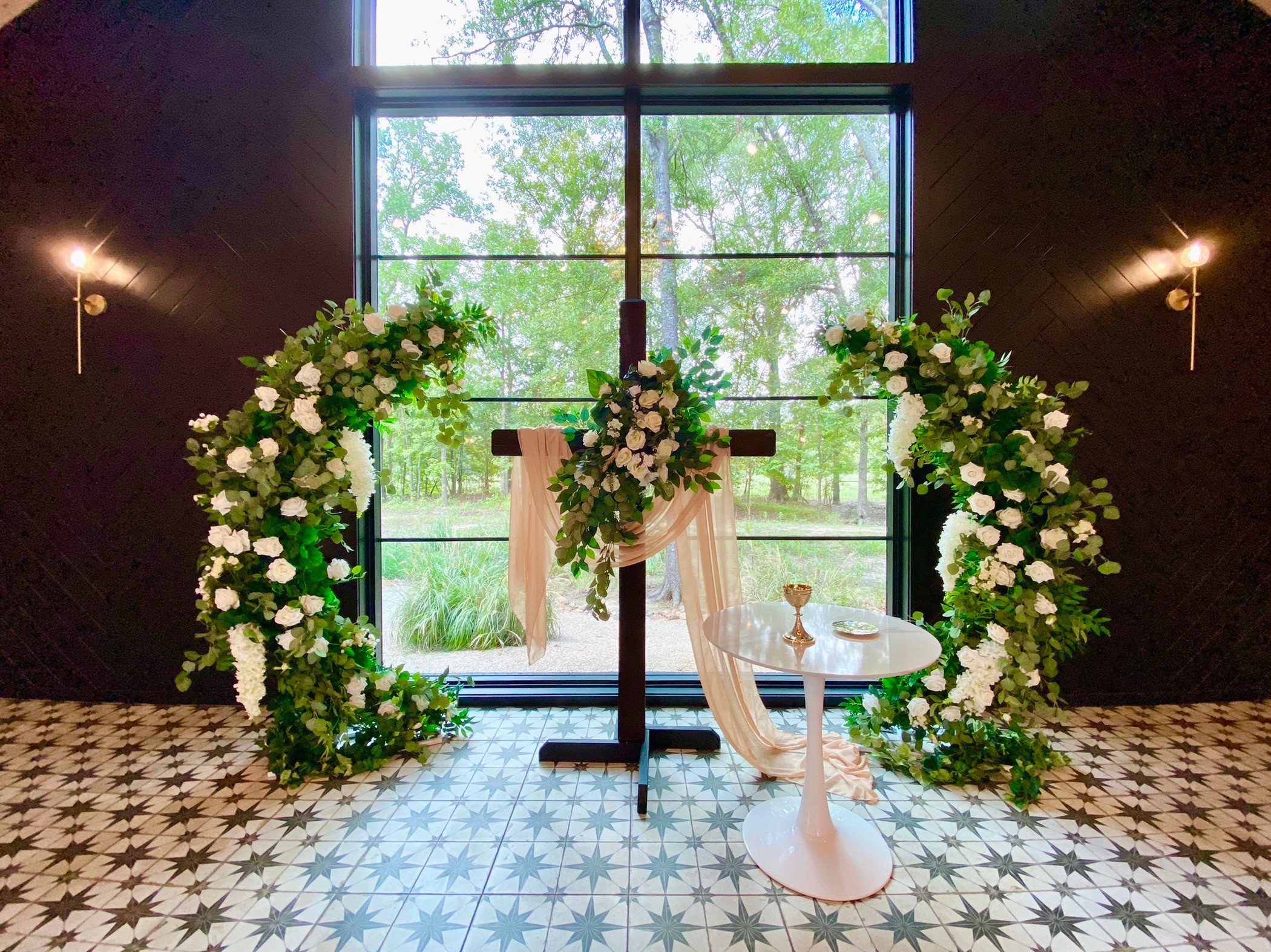 Indoor wedding setup with a cross, floral arch, and small table near large window with trees outside.