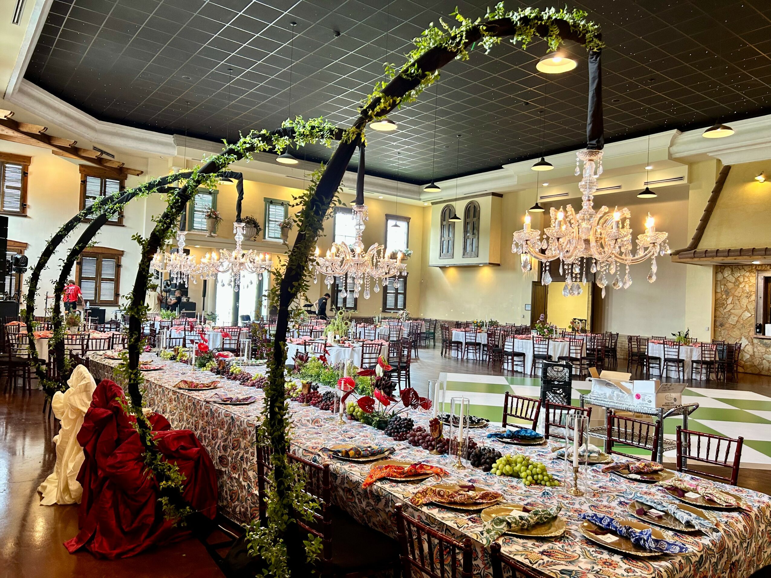 Elegant banquet hall with decorated tables, floral arrangements, chandeliers, and a floral arch, set for a celebration or event.