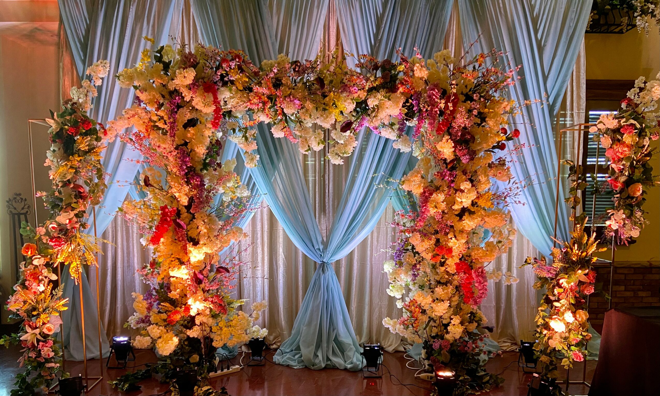 Floral arch with pink, yellow, and white flowers, blue drapes, and soft lighting, set for a celebration or wedding.